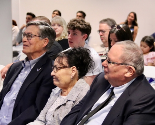 Photograph of a group of people seated indoors, likely attending a formal event or meeting. Attendees are dressed in business or semi-formal attire, with rows of chairs and a neutral background indicating a conference or presentation setting.