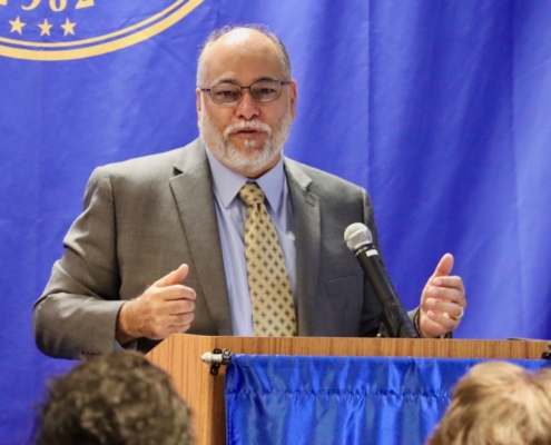 Photograph of a man in a suit and patterned tie speaking at a podium with a microphone, set against a blue backdrop featuring a partial emblem. Audience members are visible in the foreground, indicating a formal event or press conference setting.