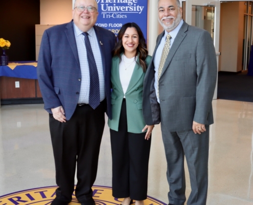 Photograph of three professionally dressed individuals standing on a Heritage University emblem inside a building lobby. A blue sign with white text reading "Heritage University Tri-Cities" is visible in the background, indicating an academic or formal event setting.