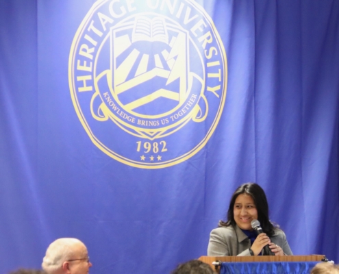Photograph of a speaker standing at a podium with a microphone in front of a blue backdrop featuring Heritage University’s emblem and motto "Knowledge Brings Us Together" along with the founding year 1982. Audience members are partially visible in the foreground, indicating a formal event or presentation setting.