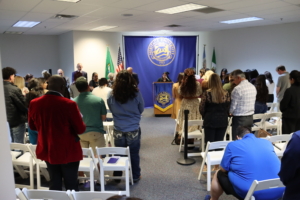 Photograph of a formal gathering in a conference room with attendees standing and facing a speaker at a podium adorned with an official seal and banners. Flags flank the podium, and white folding chairs are arranged in rows, indicating a ceremonial or official event.