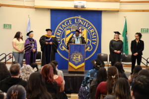 Photograph of a university graduation ceremony with faculty members in academic regalia and graduates standing on stage in front of an audience. A large blue banner with 