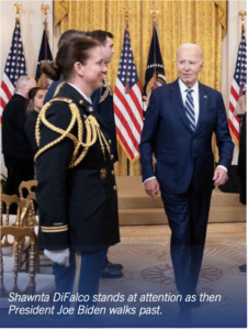 Photograph showing a formal setting with multiple American flags and gold curtains in the background. A uniformed military personnel stands at attention while a man in a suit, identified as then President Joe Biden, walks past.