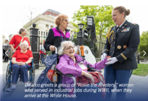 Photograph showing a military officer greeting elderly women, identified as "Rosie the Riveters," who served in industrial jobs during WWII, outside a government building. The scene includes women in wheelchairs and casual clothing, with greenery and architectural elements in the background, highlighting a moment of recognition and honor.