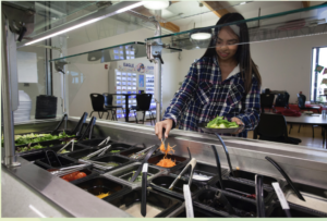 A student serves herself from a campus salad bar, adding shredded carrots to a bowl of fresh greens in a bright dining area.