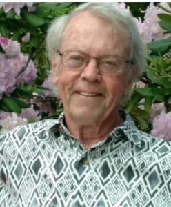 Photograph of an elderly person standing outdoors in front of blooming pink flowers and green leaves. The person wears a patterned shirt with geometric shapes in black and white.