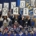 Photograph of a group of people holding large signs that collectively display "THANK YOU! $1,941,096" in a celebratory setting. The scene includes a blue and gold backdrop with a logo, and a collage of smaller photos featuring various individuals is arranged along the bottom.