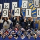 Photograph of a group of people holding large signs that collectively display "THANK YOU! $1,941,096" in a celebratory setting. The scene includes a blue and gold backdrop with a logo, and a collage of smaller photos featuring various individuals is arranged along the bottom.