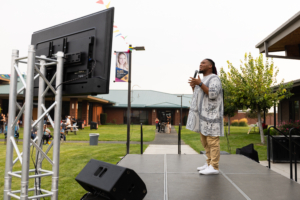 Photograph of a man speaking on an outdoor stage with a microphone, facing a large screen mounted on a metal frame. The setting includes a grassy area with buildings in the background and a few people seated or walking, suggesting a community event or presentation.