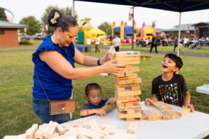 Photograph of a woman and two children playing a large outdoor Jenga game at a park event with colorful inflatable structures and people in the background. The wooden blocks have handwritten messages, and the boy on the right is laughing while the woman carefully removes a block from the tower.
