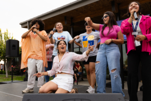 Photograph of a group of six young people performing on an outdoor stage, holding microphones and engaging with an audience. The scene features casual clothing in various colors, including a bright orange top, white shorts, and ripped jeans, with a wooden structure and greenery in the background.