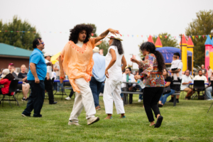 Photograph of people dancing outdoors at a festive event with colorful decorations and inflatable structures in background. Individuals wear casual and vibrant clothing, engaging in lively movement on grass with seated audience watching.