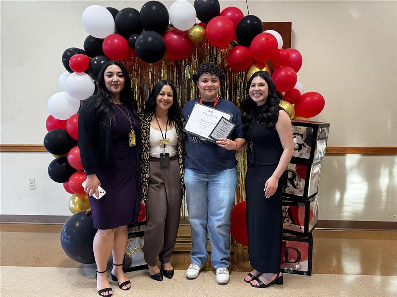 TRIO SSS 1 Photograph of four women standing in front of a decorative balloon arch with black, red, white, and gold balloons and a shimmering gold backdrop. One woman in the center holds a certificate and plaque, indicating an award or recognition event.