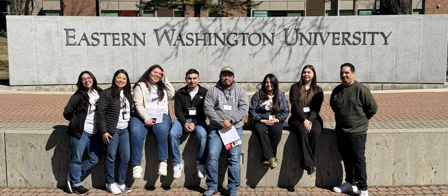 TRIO SSS 2 Photograph of eight people sitting on a concrete ledge in front of a large sign reading "Eastern Washington University" with a brick building and leafless trees in the background. The group appears relaxed and casually dressed, suggesting a campus visit or gathering on a sunny day.