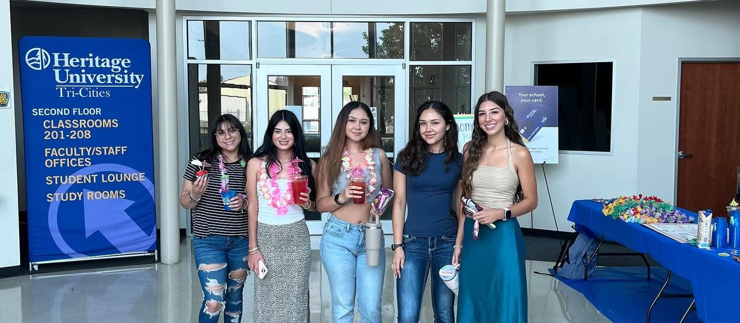 TRIO SSS 5 Photograph of five young women standing inside Ballou Hall at Heritage University, holding ice cream cones and smiling. The scene includes a large university seal on the floor, a blue banner listing campus facilities, and a table with refreshments in the background.