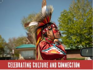 Photograph of a person dressed in traditional Native American regalia with vibrant red, black, and white feathers and geometric patterns on clothing, standing outdoors near a car mirror and greenery. Text banner below reads "Celebrating Culture and Connection," emphasizing cultural pride and community engagement.