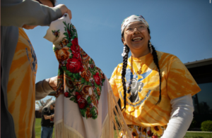 Photograph of a smiling person wearing a yellow tie-dye shirt and a white bandana, holding a colorful floral shawl with fringe. The scene takes place outdoors under a clear blue sky, suggesting a festive or cultural event.