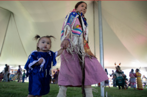 Photograph of two Native American children dressed in traditional regalia holding hands under a large event tent. The older child wears a colorful shawl and carries a walking stick, while the younger child wears a blue dress with white fringe, with other participants visible in the background.