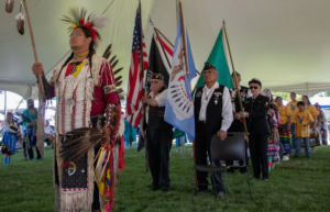 Photograph of a cultural ceremony under a large tent featuring a Native American man in traditional regalia holding a staff, followed by veterans carrying various flags including the American flag. The scene highlights respect and honor with participants dressed in ceremonial attire and a crowd observing in the background.