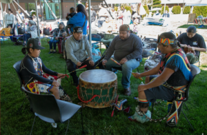 Photograph of four men seated in a circle on grass, playing a large traditional drum with drumsticks during an outdoor cultural event. Men wear colorful Native American regalia and casual clothing, with other people and tents visible in background, indicating a community gathering or celebration.