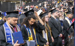 Photograph of a diverse group of graduates wearing black caps and gowns during a commencement ceremony. Graduates hold diplomas and wear colorful honor cords and stoles, with a crowded audience visible in the background.