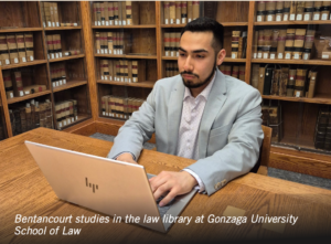 Photograph of a man studying on a laptop in a law library, surrounded by wooden shelves filled with legal books. The setting highlights a focused academic environment at Gonzaga University School of Law.