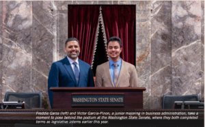 Photograph of two men standing behind a podium inside Washington State Senate chamber, with marble walls and a red velvet curtain in the background. The men, dressed in suits, are Freddie Garza and Victor Garcia-Pinon, who completed internships as legislative interns at the Senate.