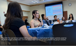 Photograph of a seminar room with students seated around tables covered in blue tablecloths, engaging in a discussion. A female student holding a microphone speaks while others listen, with a presentation slide titled "Questions for Small Group Exercise" visible on a screen in the background.