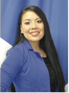 Photograph of a smiling woman with long black hair wearing a blue cardigan over a black top, standing against a blue and white background. The image highlights her friendly expression and professional appearance.