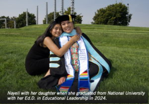 Photograph of a graduate in academic regalia sitting on grass with her daughter hugging her, celebrating completion of an Ed.D. in Educational Leadership in 2024. The graduate wears a black cap and gown with colorful stoles and cords, set against a background of green hills and trees under clear sky.