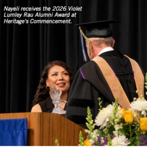 Photograph of a woman named Nayeli receiving the 2026 Violet Lumley Rau Alumni Award during Heritage's commencement ceremony. The scene features Nayeli smiling at a man in academic regalia, with a podium, a clear eagle-shaped award, and a floral arrangement with white, yellow, and purple flowers in the foreground.