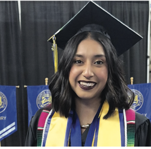 Photograph of a female graduate wearing a black cap and gown with a yellow stole and multiple honor cords, smiling in front of blue banners with university logos. The setting suggests a graduation ceremony celebrating academic achievement and honors.