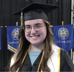 Photograph of a smiling graduate wearing a black cap and gown with a gold honor cord and white stole, standing in front of blue banners labeled "Mathematics" and "Psychology" from Heritage University. The setting highlights an academic achievement ceremony, emphasizing fields of study and celebratory atmosphere.