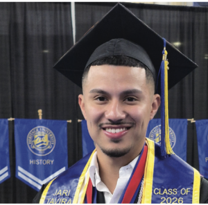 Photograph of a smiling male graduate wearing a black cap with a yellow tassel and a blue stole with gold trim. Background features blue banners with university logos and department names, highlighting a graduation ceremony for class of 2026.