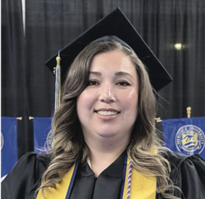 Photograph of a female graduate wearing a black cap and gown with a yellow stole and honor cords, smiling at a graduation ceremony. Blue banners with university logos are visible in the background, indicating an academic achievement celebration.