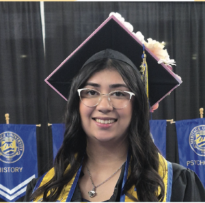 Photograph of a young woman wearing a decorated graduation cap with flowers and a tassel, along with a black gown and yellow and blue stole. Blue banners with university logos and academic disciplines like History and Psychology are visible in the background, indicating a graduation ceremony setting.