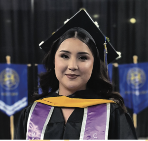 Photograph of a female graduate wearing a black cap and gown with a purple and gold stole, standing in front of blue banners. The setting suggests a formal graduation ceremony, highlighting academic achievement and celebration.