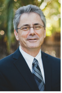 Photograph of a middle-aged man wearing glasses, a dark suit, white shirt, and patterned tie, standing outdoors with blurred greenery and sunlight in the background. The image focuses on the man's face and upper body, highlighting a professional and approachable appearance.