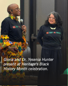 Photograph of two women speaking at Heritage's Black History Month celebration, both wearing black shirts with "WE CAN'T BE ERASED" in red, green, and white text. One woman holds a microphone and wears colorful patterned clothing underneath her shirt, while the other smiles and stands with hands behind her back.