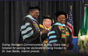 Photograph of a commencement ceremony showing three individuals in academic regalia, with one woman being hooded by a man. The scene includes colorful robes with blue and gold accents, an American flag, and a green flag in the background, highlighting the honor of earning a doctorate during Heritage's May event.