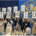 Photograph of a group of people standing in front of a blue curtain holding large signs that spell out "THANK YOU! $1,941,096" at an indoor event. The scene includes seated attendees at round tables with floral centerpieces, indicating a celebratory fundraising or donation announcement.