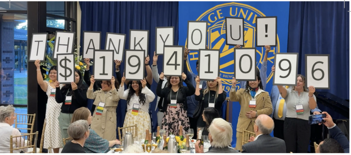 Photograph of a group of people standing in front of a blue curtain holding large signs that spell out "THANK YOU! $1,941,096" at an indoor event. The scene includes seated attendees at round tables with floral centerpieces, indicating a celebratory fundraising or donation announcement.