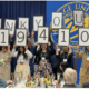 Photograph of a group of people standing in front of a blue curtain holding large signs that spell out "THANK YOU! $1,941,096" at an indoor event. The scene includes seated attendees at round tables with floral centerpieces, indicating a celebratory fundraising or donation announcement.