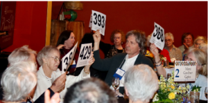 Photograph of a group of people participating in an auction, holding up numbered paddles to place bids. The setting includes a table with floral centerpieces and a sign labeled "Mimi Gardner Gates," indicating a formal or charity event.