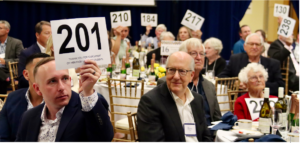 Photograph of a fundraising auction event showing attendees seated at tables holding numbered bidding paddles. Key details include paddle numbers such as 201, 210, and 217, with tables set with wine bottles, glasses, and plates, indicating an organized social gathering.