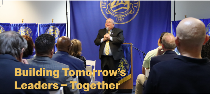 Photograph of a speaker addressing an audience at a leadership event, with a blue backdrop displaying a seal and the year 1982. Text overlay in yellow reads "Building Tomorrow's Leaders – Together," emphasizing collaboration and future leadership development.