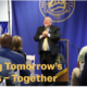 Photograph of a speaker addressing an audience at a leadership event, with a blue backdrop displaying a seal and the year 1982. Text overlay in yellow reads "Building Tomorrow's Leaders – Together," emphasizing collaboration and future leadership development.