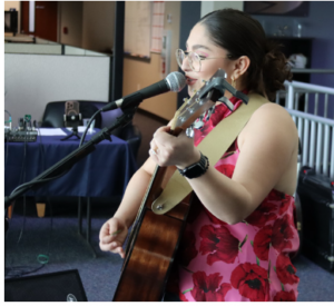 Photograph of a young woman playing an acoustic guitar and singing into a microphone during a live performance. She wears glasses and a red floral dress, with audio equipment and a mixer visible in the background.