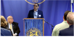 Photograph of a man speaking at a podium with a Heritage University banner and blue backdrop featuring the university's seal and founding year, 1982. Audience members are seated facing the speaker, indicating a formal event or presentation.