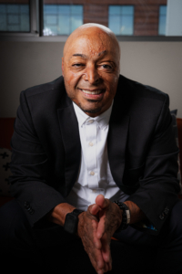 Portrait photograph of a smiling bald Hispanic man seated indoors, wearing a black suit jacket and white shirt. Soft window light and blurred office-like background create a professional, approachable presentation.